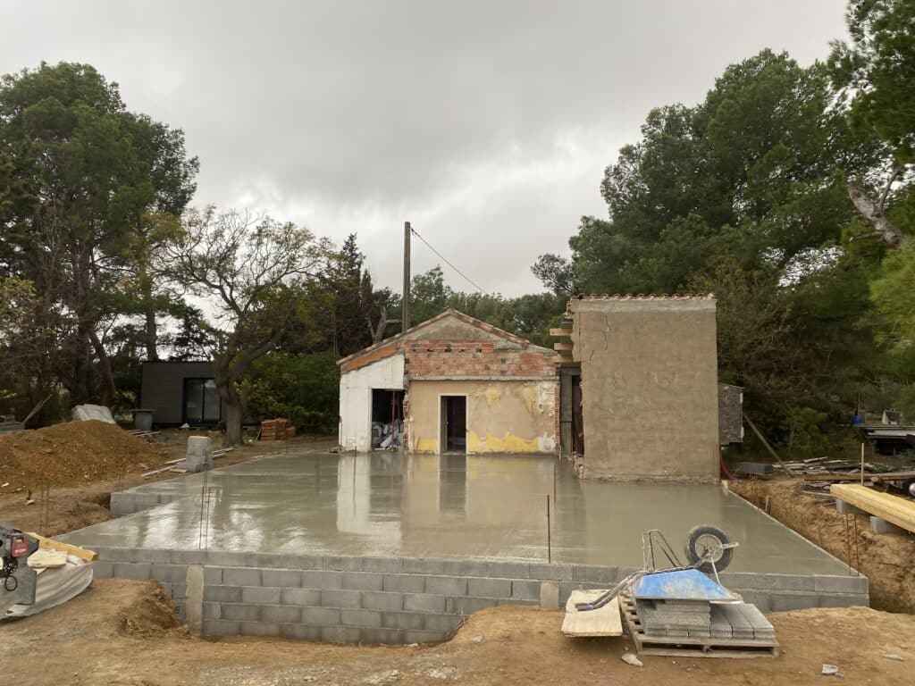 Construction de dalle béton pour rénovation maison Dalle béton lissée devant une maison en rénovation, chantier avec brouette sous un ciel couvert.