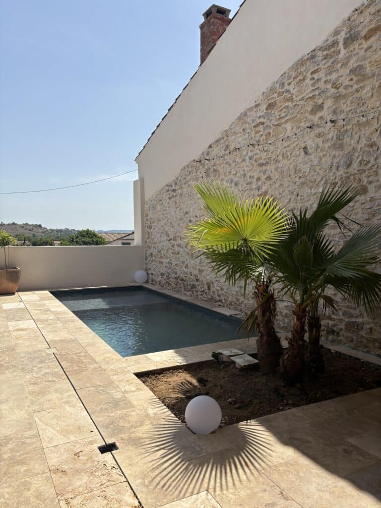 Piscine moderne avec terrasse en pierre et palmier Petite piscine moderne avec terrasse en pierre, mur de pierre sèche, palmier et luminaires boules sous le soleil.