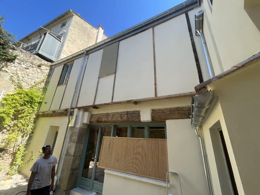 Rénovation de Façade Ancienne : Maison à Colombages Homme contemplant la rénovation d'une façade ancienne à colombages avec gouttières en zinc.